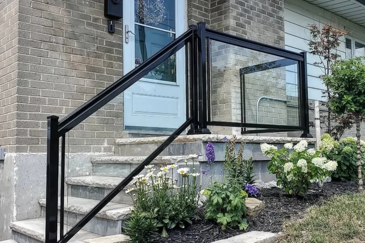 Clear glass railing in a black aluminum frame beside stone front steps and garden planting